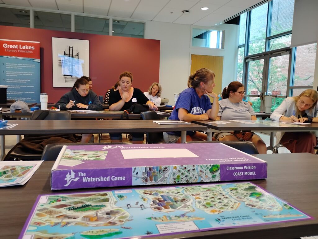 Educators work in small groups playing the Watershed game. The Watershed Game Coastal Model frames the bottom of the image.