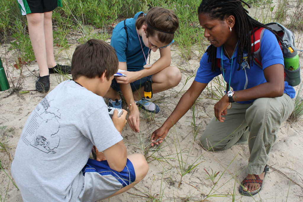 A park ranger shows 2 students a plant in a dune