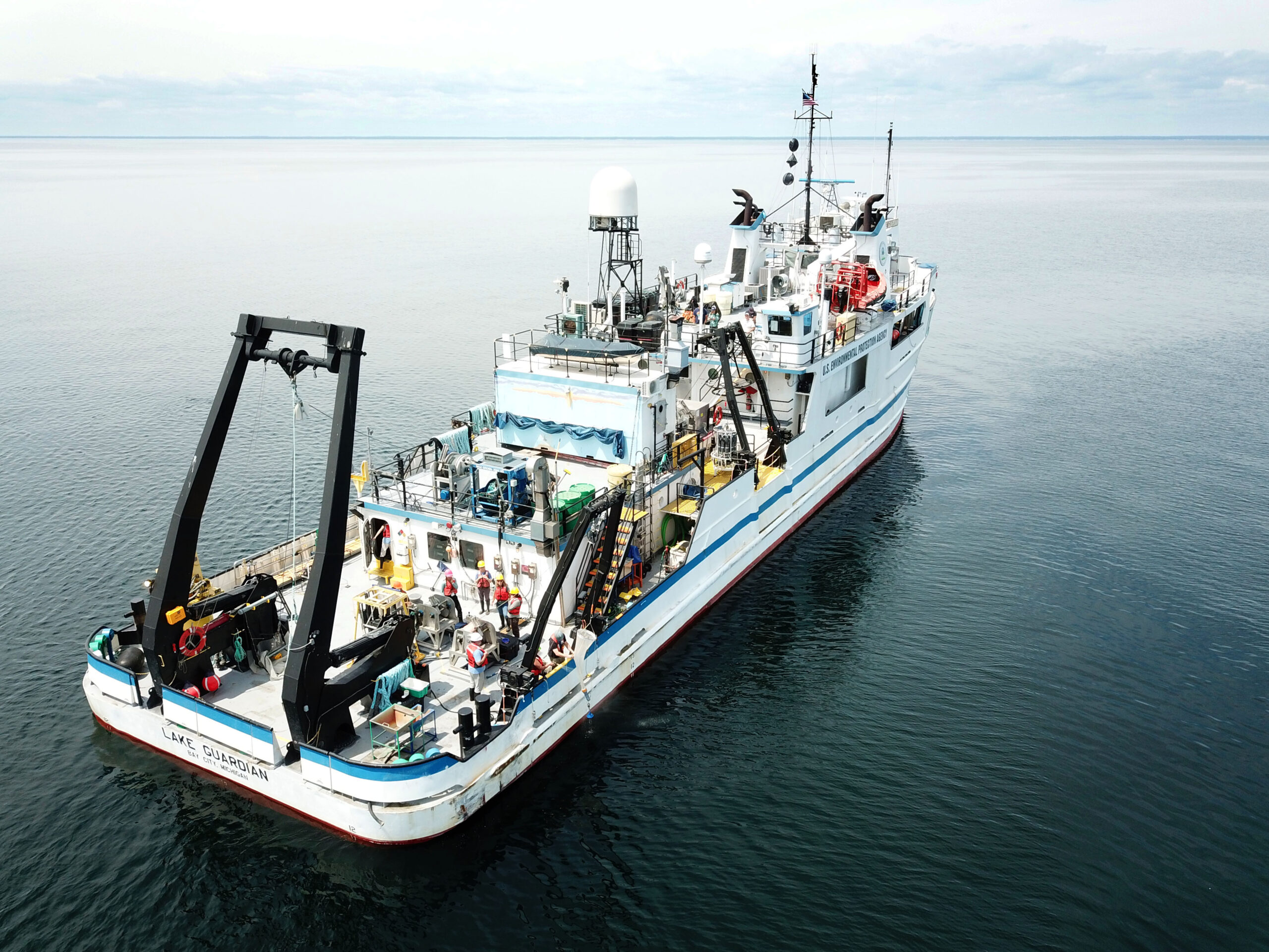 Aerial drone image of R/V Lake Guardian on Lake Michigan taken from stern perspective. Large A-frame with pully system for lowering and raising equipment is visible on stern fantail deck. Navigation and meteorological sensors extend from the roofs of the hotel deck and pilot house.