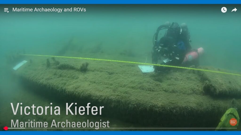 Dressed in her scuba gear, Victoria Kiefer, maritime archaeologist, Wisconsin Historical Society hovers above a shipwreck. The wreck has a fuzzy appearance due to the greenish algae growing on its surface. A yellow measuring tape floats above the length of the ship. A white dive slate, upon which measurements and drawings are being recorded, is another important piece of equipment.