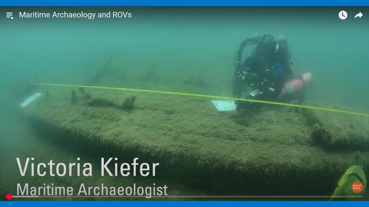 Dressed in her scuba gear, Victoria Kiefer, maritime archaeologist, Wisconsin Historical Society hovers above a shipwreck. The wreck has a fuzzy appearance due to the greenish algae growing on its surface. A yellow measuring tape floats above the length of the ship. A white dive slate, upon which measurements and drawings are being recorded, is another important piece of equipment.