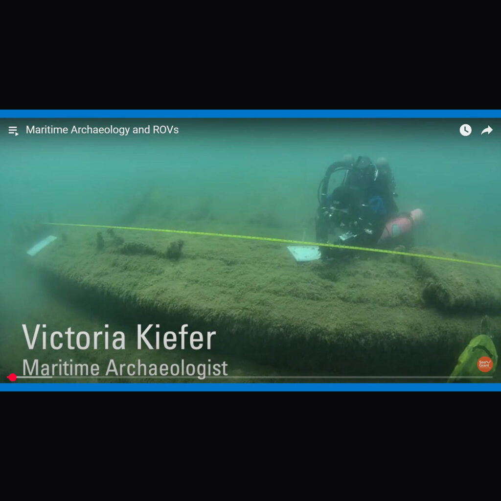 Dressed in her scuba gear, Victoria Kiefer, maritime archaeologist, Wisconsin Historical Society hovers above a shipwreck. The wreck has a fuzzy appearance due to the greenish algae growing on its surface. A yellow measuring tape floats above the length of the ship. A white dive slate, upon which measurements and drawings are being recorded, is another important piece of equipment.