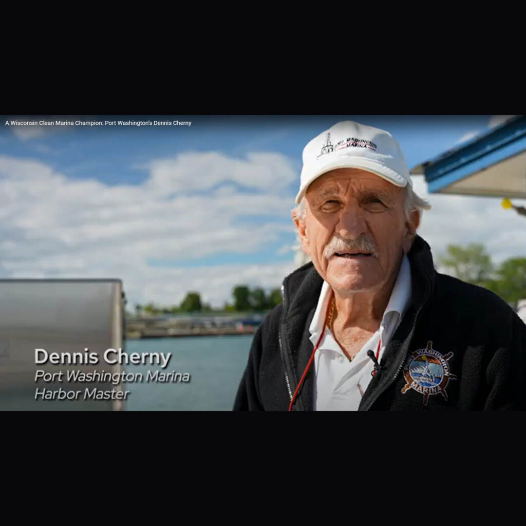 Dennis Cherny Port Washington Harbor Master wears a white baseball cap and black fleece jacket each with the words "Port Washington Marina" on it. He stands on a dock within the marina.