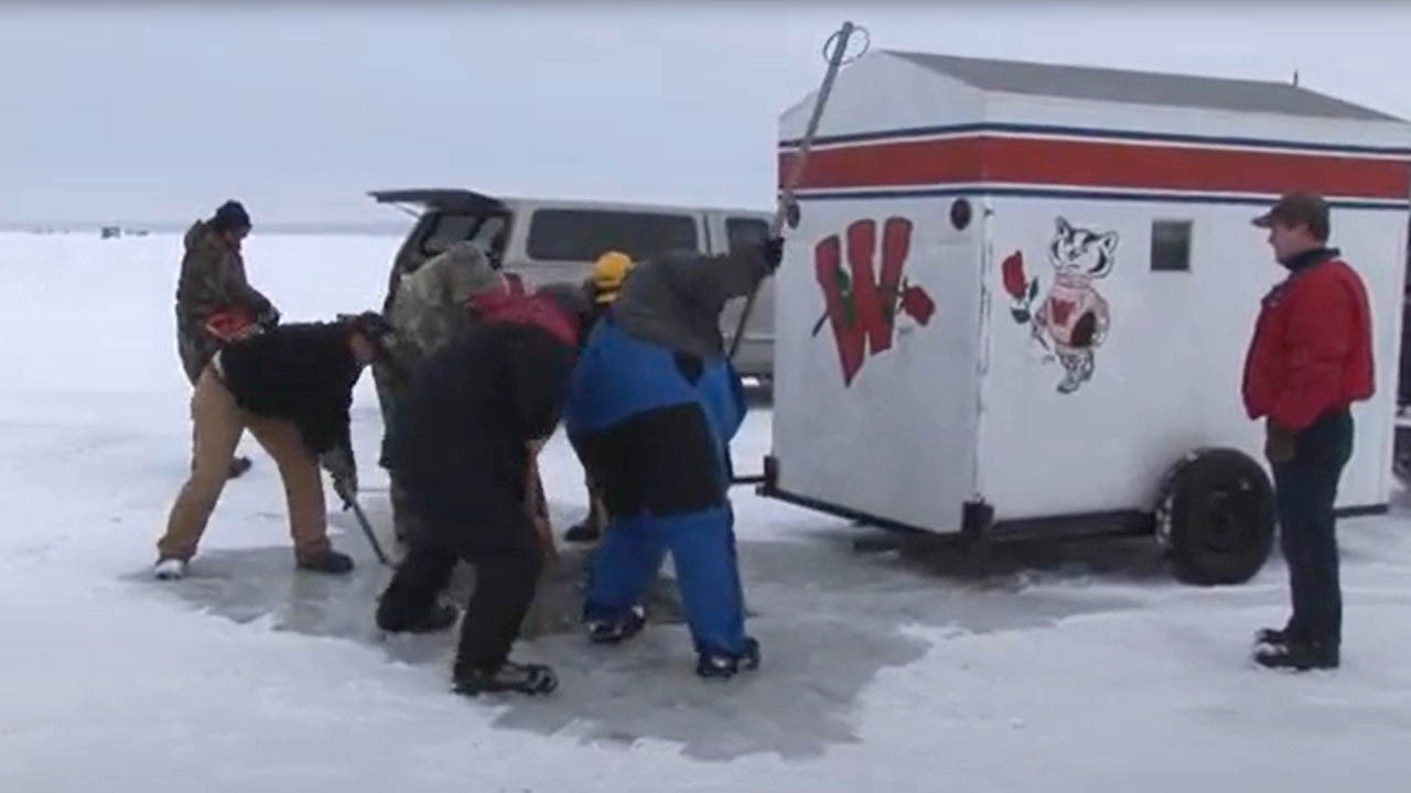 A group of five people dressed in warm clothing stand around a rectangular hole in the ice. Several people have poles to manipulate the block of ice that has been cut away from the hole. Two additional people stand nearby looking on. An ice shanty decorated with the University of Wisconsin "W" and Bucky Badger logo sits on a trailer nearby. A vehicle is parked along-side the ice-shanty.