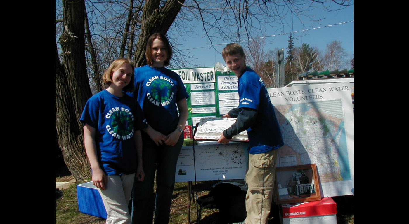Students Maree, Janelle, and Luke, in the original Clean Boats, Clean Waters shirts, pose at their outdoor educational booth.